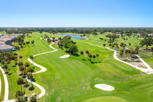 Elevated drone photograph of a lush green golf course featuring manicured fairways, sand traps, and surrounding residential community under a clear blue sky.
