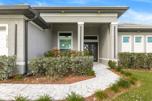 A wide-angle, high-definition (HDR) photograph of a modern gray house with a white garage door, paved driveway, and palm trees under a bright blue sky.
