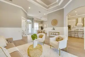 Professional interior photograph by Realty Media 360 of a bright living room featuring white armchairs, a gold accent table, and an open layout leading into the kitchen area.